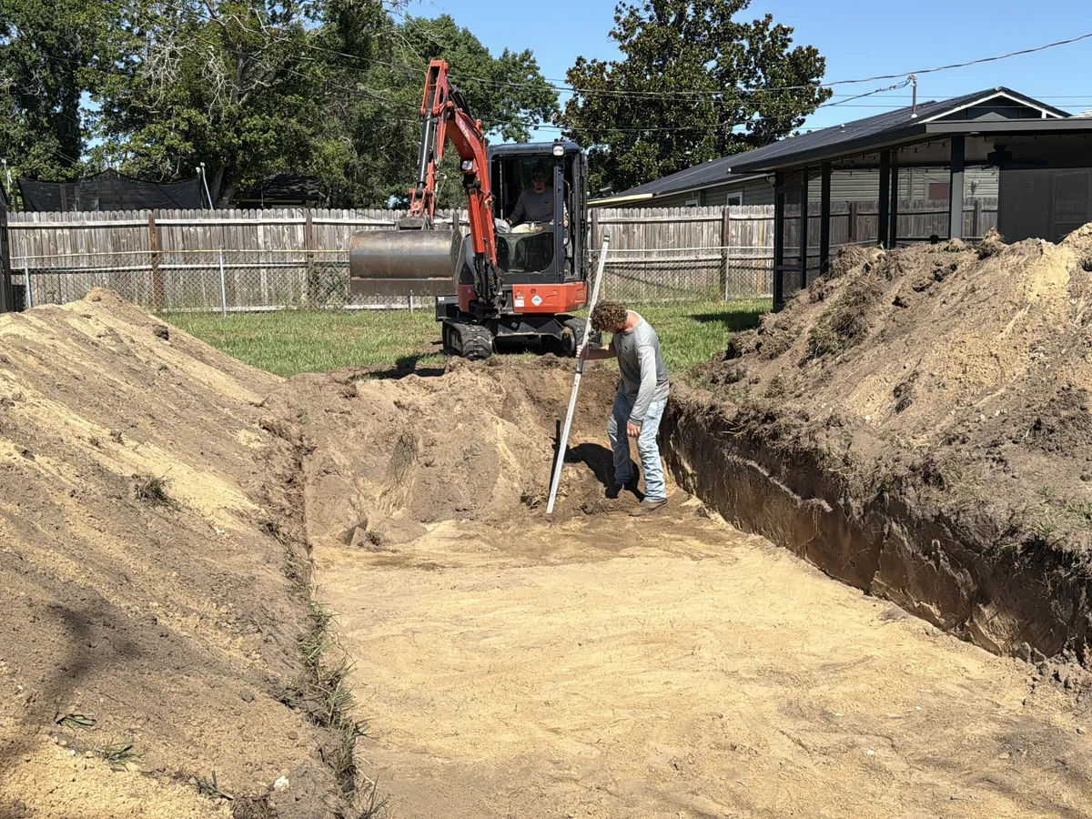 A long drain field trench dug through sandy soil behind a house, with a mini excavator at the far end and a worker checking the trench bottom with a grade rod