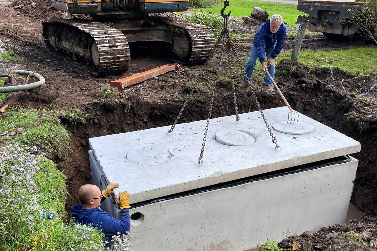 A new concrete septic tank being lowered into the dig on chains from an excavator, with a worker guiding it from the trench
