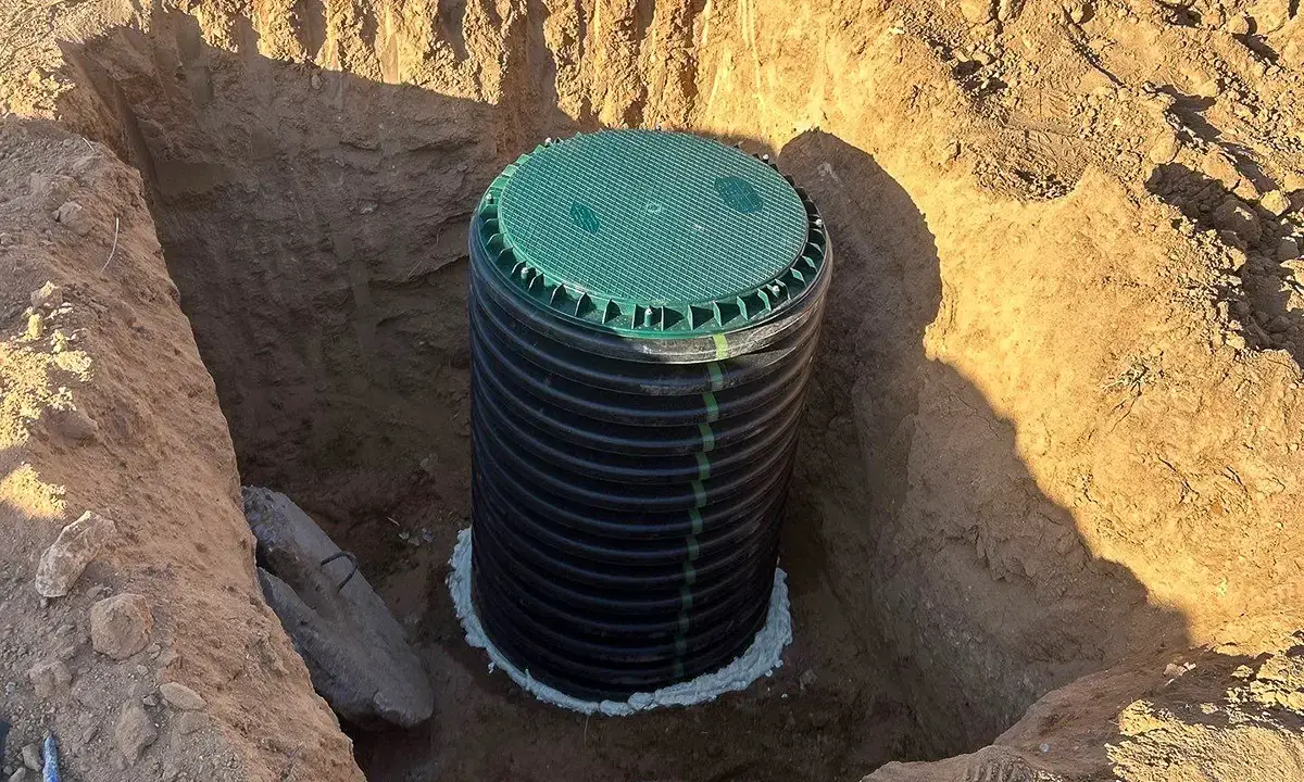 A new septic tank riser with a green lid set on a buried tank in an open excavation, the old concrete lid lying beside it